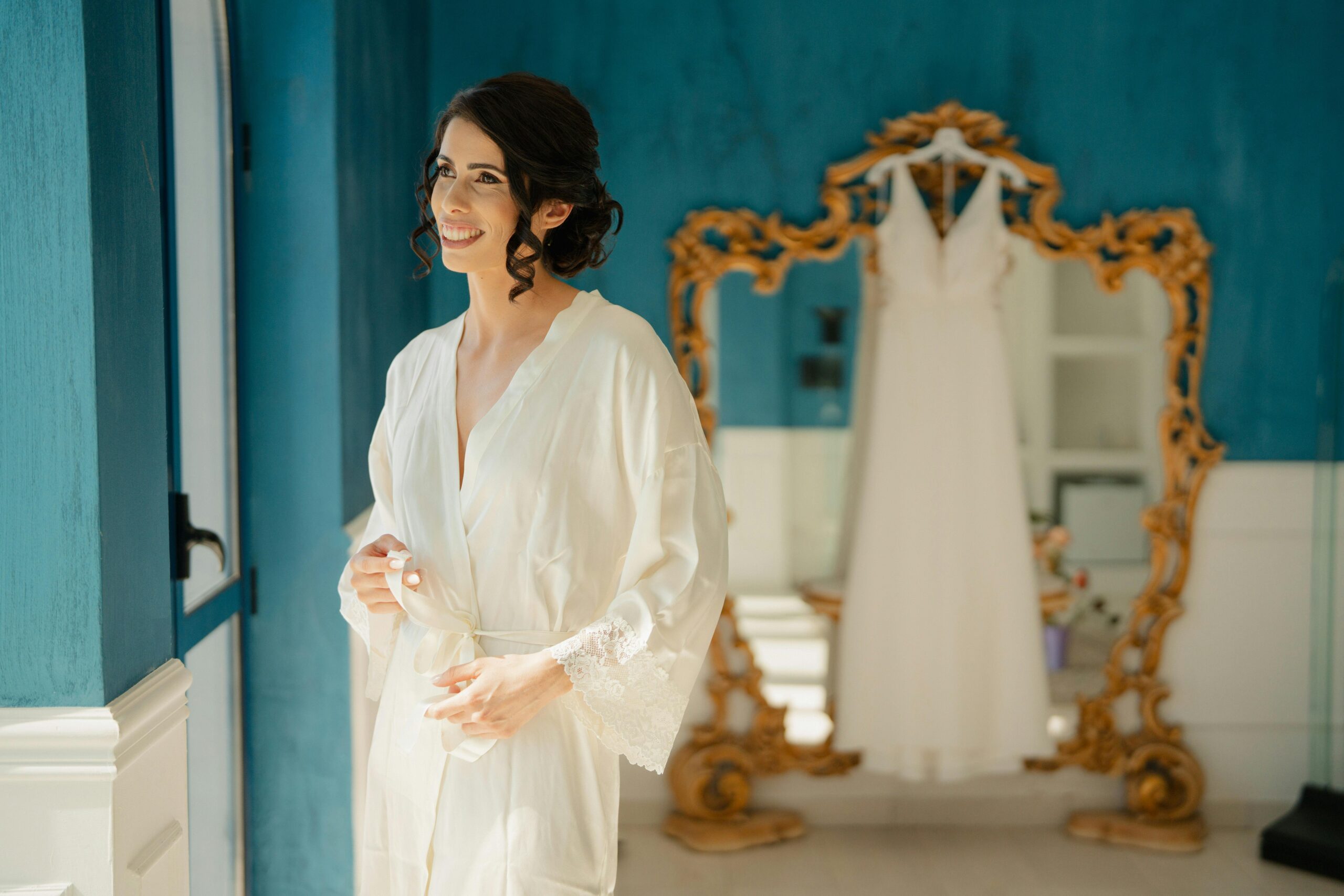 A bride enjoys a serene moment in a blue room with her wedding dress in the background.